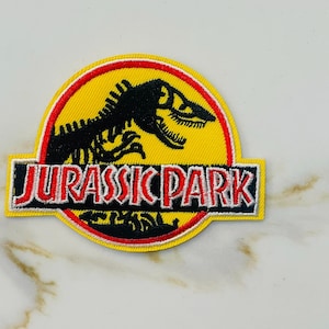 May include: A yellow, red, and white embroidered patch with the Jurassic Park logo. The logo features a dinosaur skeleton and the words "Jurassic Park".