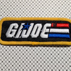 May include: Embroidered patch with the text "G.I. JOE" in white, featuring a blue star and red, white, and blue stripes. The patch has a black background with a yellow border. The patch is rectangular.