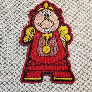 May include: Embroidered patch featuring a cartoon character in a red outfit with yellow accents. The character has a clock face and a smiling expression. The patch is outlined in black and has a decorative border.