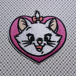 May include: Embroidered patch featuring a white cat face with black whiskers and eyes, set within a pink heart-shaped frame. A small pink bow sits atop the cat's head. The patch is designed for application on clothing or accessories.