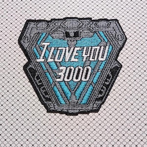 May include: Embroidered patch in a shield shape with a teal and gray design. The text "I LOVE YOU 3000" is embroidered in white. The patch has a black border and is on a patterned background.