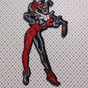 May include: Embroidered patch featuring a stylized character in a red, black, and white color scheme. The patch depicts a figure in a dynamic pose, with a harlequin-inspired design. The background is a repeating pattern of small, interconnected shapes.