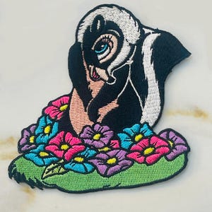 May include: An embroidered iron-on patch featuring a cartoon skunk with a flower crown. The skunk is black and white with a pink nose and blue eyes. The flowers are pink, blue, and purple.