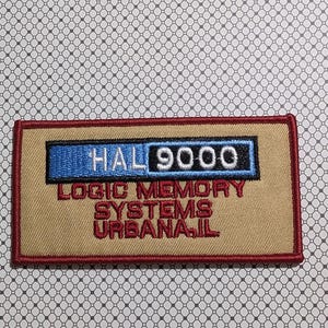 May include: Embroidered patch with the text "HAL 9000" in white, blue, and black, above "LOGIC MEMORY SYSTEMS URBANA, IL" in red. The patch has a tan background with a red border.