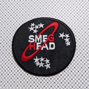 May include: A black, circular patch with the text "SMEG HEAD" in white and red, encircled by a red loop. White star-shaped designs are scattered around the text and loop. The patch is on a patterned background.