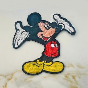May include: An embroidered iron-on patch featuring Mickey Mouse in a classic pose with his signature red shorts and yellow shoes.