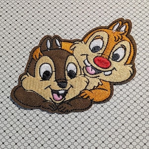 May include: Embroidered patch featuring Chip and Dale, the Disney chipmunk duo. Chip is brown, and Dale is tan with a red nose. Both have large, expressive eyes and are smiling. The patch is on a patterned background.