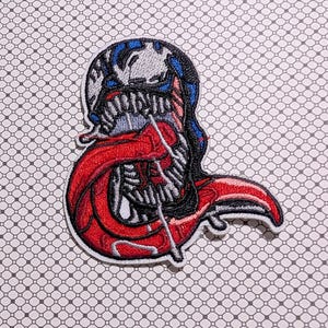 May include: Embroidered patch featuring a stylized monster design with a red tongue, sharp teeth, and a black, white, and blue head. The patch has a white border and is set against a patterned background.