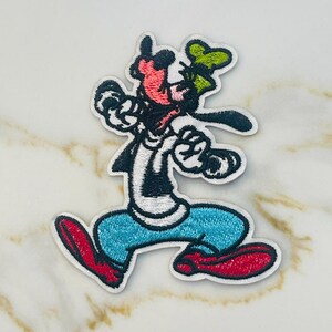 May include: An embroidered iron-on patch featuring a cartoon image of Goofy in a blue shirt, red shoes, and a green hat. He is walking with his arms outstretched.