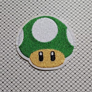 May include: Embroidered patch featuring a green mushroom with a white top and a tan base. The mushroom has two black oval-shaped eyes. The patch is likely for clothing or accessories.