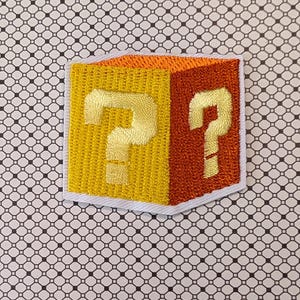 May include: Embroidered patch featuring a yellow and orange cube with two gold question marks. The patch has a white border and is set against a patterned background. The design is reminiscent of a classic video game.