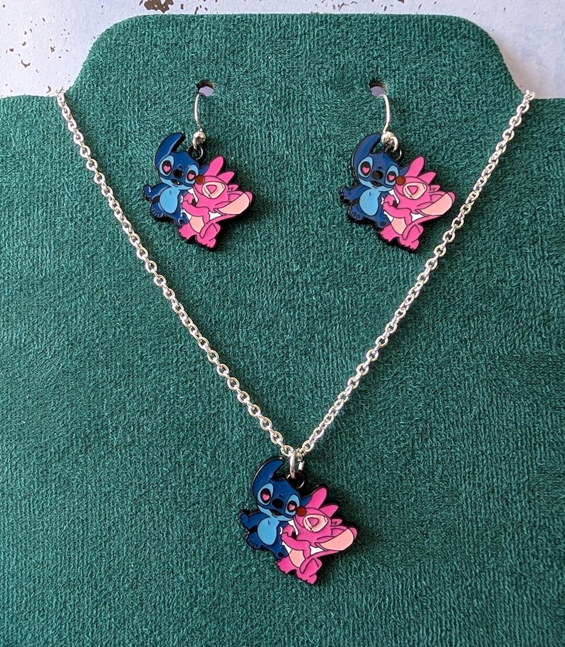May include: A silver-toned necklace and earring set featuring blue and pink cartoon character pendants. The pendants depict two characters embracing. The earrings hang from silver hooks. The necklace has a delicate chain.