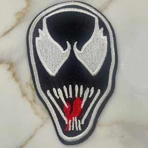 May include: Black and white embroidered patch of Venom's face with a red tongue. The patch is a popular character from Marvel Comics.