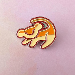 May include: Enamel pin featuring the silhouette of a lion cub in shades of orange, yellow, and red, with a silver outline. The pin is set against a soft pink background.