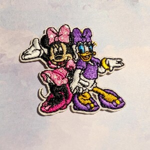 May include: Embroidered patch featuring Minnie Mouse and Daisy Duck. Minnie Mouse is wearing a pink dress and bow, while Daisy Duck is in a purple outfit. Both characters are smiling and waving, with white gloves.
