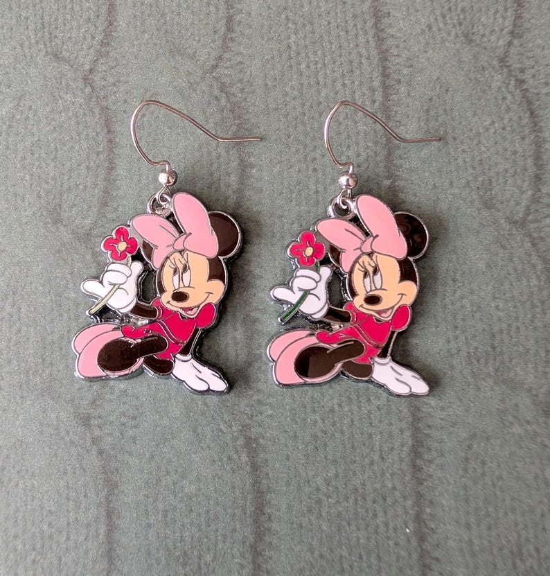 K&ouml;nnte beinhalten: Ein Paar silberfarbene Minnie Mouse-Ohrringe. Jeder Ohrring zeigt eine Cartoon-Figur von Minnie Mouse in einem rosa Kleid und einer Schleife, die eine rote Blume h&auml;lt. Die Ohrringe haben einen Hakenverschluss.