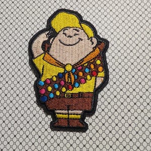 May include: Embroidered patch featuring a cartoon character in a yellow shirt, brown shorts, and a yellow hat. The character has a brown sash with colorful circular badges. The patch has a black border and is on a patterned background.