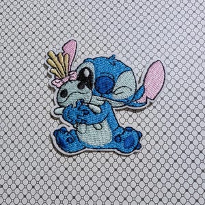 May include: Embroidered patch featuring a blue cartoon character with large ears and a playful expression. The character is holding a smaller version of itself. The patch has pink, yellow, and white accents, and a white border.