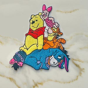 May include: An embroidered patch featuring Winnie the Pooh, Piglet, Tigger, and Eeyore. The characters are stacked on top of each other, with Eeyore on the bottom and Winnie the Pooh on top. The patch is made of felt and has a colorful design.