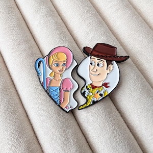 May include: Two-piece enamel pin set featuring cartoon characters Bo Peep and Woody from Toy Story. Bo Peep is in a pink dress and hat, holding a blue shepherd's crook. Woody is in a cowboy hat and vest. The pins are in the shape of a broken heart.