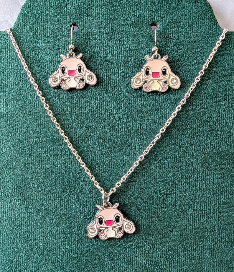 May include: A silver-toned jewelry set featuring a pendant necklace and matching earrings. Each piece showcases a pink and white cartoon character design with black eyes, a red mouth, and clear crystal accents. The set is displayed on a green fabric.