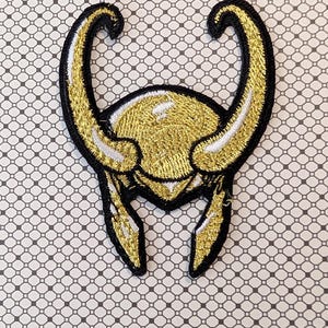 May include: Embroidered patch featuring a gold and black horned helmet design. The patch has a gold center with black outlining and white accents. The patch is likely for clothing or accessories.