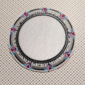 May include: Circular embroidered patch with a silver center and a black border. The border features stylized text and red and blue accents. The patch is on a patterned background.