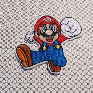 May include: Embroidered patch featuring the character Mario from the Super Mario Bros. video game series. Mario is depicted in his signature red hat, blue overalls, and brown shoes, with a white glove raised in a jumping pose.