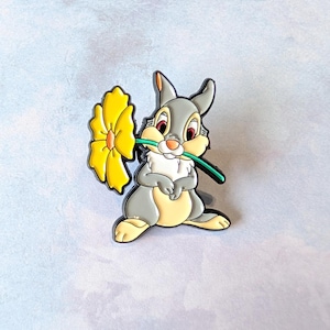 May include: A gray and yellow cartoon bunny holding a yellow flower in its mouth. The bunny has red eyes and a white chest. The flower has a green stem. The image is on a light blue background.