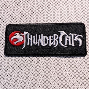 May include: Black rectangular patch with the words "THUNDERCATS" embroidered in white. The patch features a red circle with a white cat-like figure. The patch is on a patterned background.