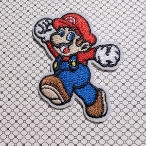 May include: Embroidered patch featuring a cartoon image of Mario in a running pose. Mario is wearing a red hat, blue overalls, and brown shoes. The patch has a white border and is set against a patterned background.