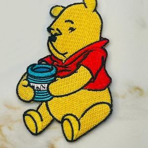 May include: Embroidered iron-on patch featuring Winnie the Pooh, a beloved cartoon bear, sitting and holding a blue pot. Winnie is wearing a red shirt and has a yellow body.