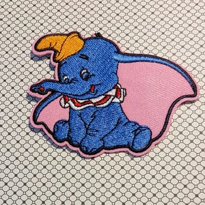May include: Embroidered patch featuring Dumbo the elephant from Disney. The patch shows Dumbo in blue with a yellow hat, pink ears, and a red and white collar. The patch has a pink border.