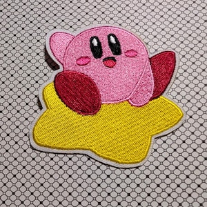 May include: Embroidered patch featuring a pink character with large black eyes, small pink cheeks, and a red mouth. The character is on a yellow star-shaped base with a white border. The patch is likely for clothing or accessories.