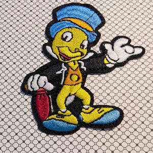 May include: Embroidered patch featuring a cartoon character in a blue top hat and black jacket. The character has a yellow face, orange vest, and blue shoes. It is holding a red bag and has white gloves.