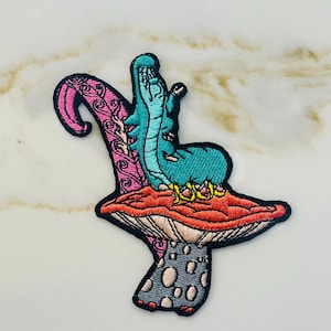 May include: An embroidered iron-on patch featuring a blue caterpillar sitting on a red and pink mushroom. The mushroom has a gray stem with white polka dots.