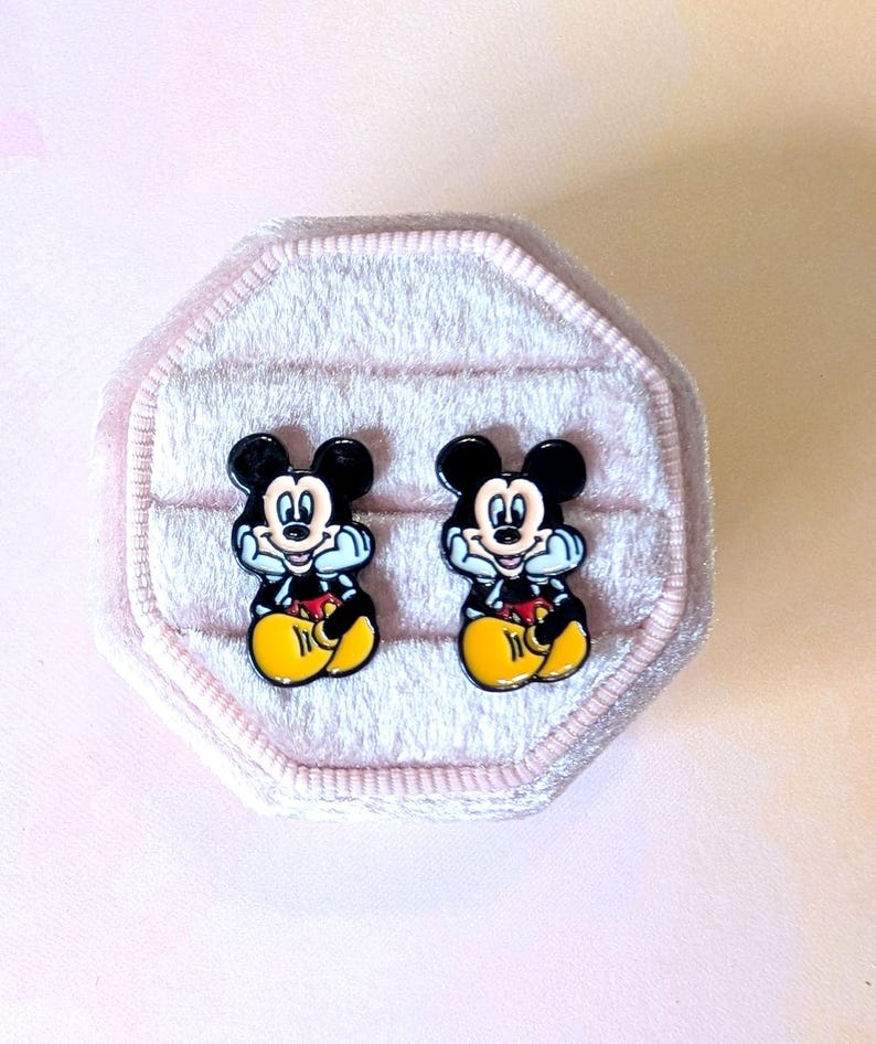 Puede incluir: Un par de pendientes de bot&oacute;n de Mickey Mouse. Cada pendiente presenta una silueta negra de la cabeza y la cara de Mickey, con una boca blanca y detalles azules. Tambi&eacute;n se ven los pantalones cortos amarillos y los guantes rojos de Mickey. Los pendientes se presentan en una caja de joyer&iacute;a de terciopelo rosa.