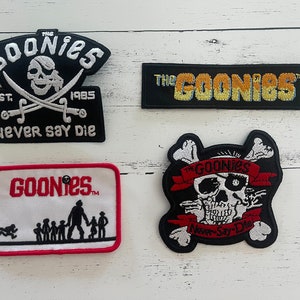 May include: Four embroidered patches featuring the Goonies logo. The patches are black, white, red, and yellow. The text on the patches reads "The Goonies", "Never Say Die", and "Goonies".
