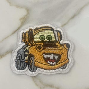 May include: A yellow embroidered patch of a cartoon tow truck with a toothy grin and green eyes. The tow truck is on a white background.