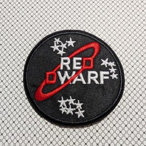 May include: A circular black patch with the words "RED DWARF" in white, with a red swirl and star clusters. The patch is embroidered and has a black border. The background is a white and gray patterned surface.