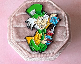 May include: A colorful Mad Hatter enamel pin. The pin features the Mad Hatter in a green hat with a white label, a yellow jacket, and a green bow tie. He is holding a blue teapot and a stack of red and blue teacups.