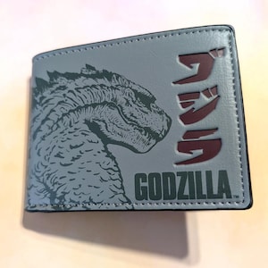 May include: A gray bi-fold wallet featuring a Godzilla design. The wallet has a Godzilla illustration on the front, along with the word "GODZILLA" and Japanese text in a dark red color.