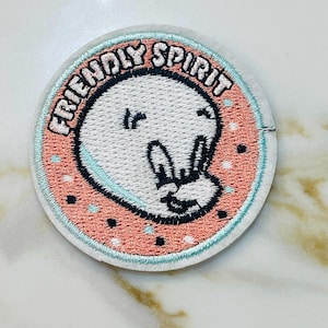 May include: A round, embroidered patch with a peach background and a white outline. The patch features a white cartoon ghost with a peace sign hand gesture and the text "FRIENDLY SPIRIT" in a white cursive font.