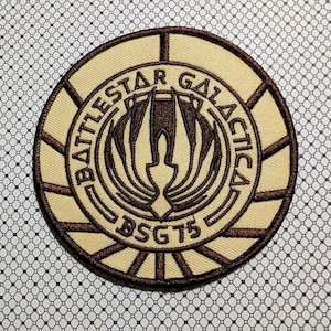 May include: A circular patch with the words "Battlestar Galactica" in a brown font, with a brown emblem in the center. The patch has a tan background with brown border and accents. The text "BSG75" is at the bottom.