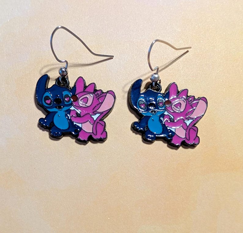 Op de afbeelding: Een paar bungelende oorbellen met blauwe en roze stripfiguren. De oorbellen hebben zilverkleurige haken. De karakters omarmen elkaar. De oorbellen liggen op een lichtbruin oppervlak.