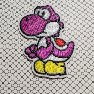 May include: Embroidered patch featuring a purple Yoshi character with white accents, a red saddle, and yellow shoes. The patch has a white border and is designed in a cartoon style. This patch is suitable for clothing or accessories.
