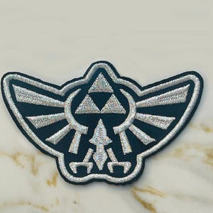 May include: A silver and black embroidered patch featuring the iconic Triforce symbol from the Legend of Zelda video game series. The Triforce is surrounded by a stylized winged design.