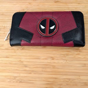May include: A red and black faux leather wallet with a metal Deadpool logo. The wallet has a zipper closure.