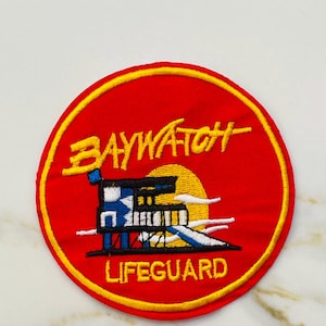 May include: Embroidered patch featuring the text "BAYWATCH" in yellow, arched above a lifeguard tower graphic. The patch is circular, with a red background and a yellow border. The word "LIFEGUARD" is written in yellow below the tower.