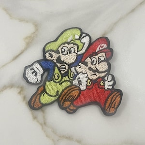 May include: Embroidered patch featuring the characters Mario and Luigi from the Super Mario Bros. video game. Mario is in a red hat and overalls, while Luigi wears a green hat and overalls. The patch has a black border and is on a white marble surface.